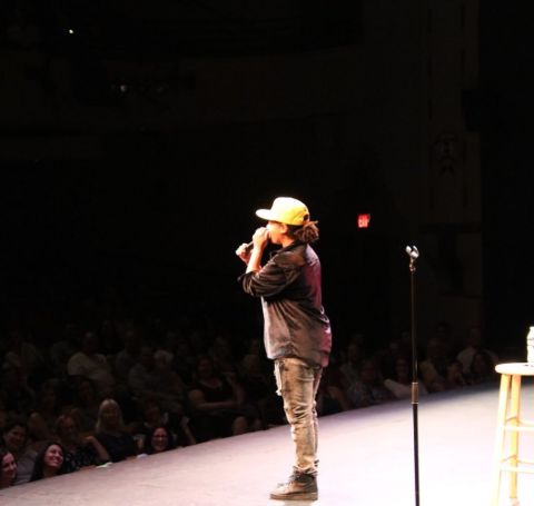 Chaunte Wayans wearing a cap and mike in her hand performs stand-up comedy in front of audience.