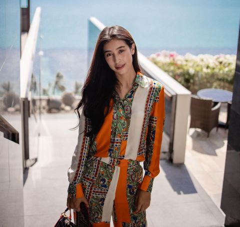 Chalida Vijitvongthong in a orange-white designer cloth in front of an ocean.