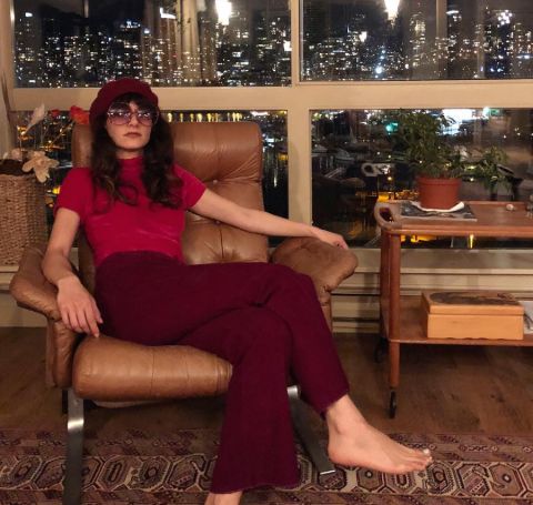 Sheila Vand in a red cap, tshirt and trouser posing at a sofa. 