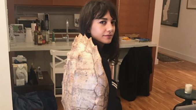 Sheila Vand looks at the camera in a turtle shell on her back.