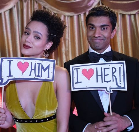 Nikesh Patel holding a I love her card to Nathalie Emmanuel in yellow dress, who holds I love Him card.