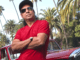 Bikram Choudhury posing in a red t-shirt and black cap.