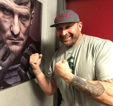 Tony Mandarich in a grey t-shirt poses beside a UFC poster.