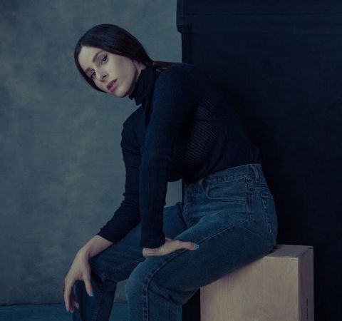 Sasha Spielberg in a black top and blue jeans poses for a picture.